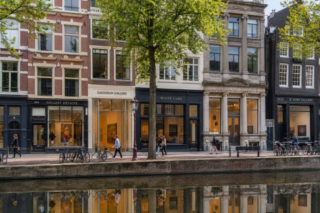 Street view of five art galleries in Amsterdam with historic buildings, canal, bicycles, and people walking nearby.