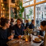 Cozy restaurant interior in Amsterdam with diners enjoying meals near large windows overlooking a street with canal houses and bicycles.