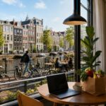 View of Amsterdam canal with traditional houses, bicycles parked along the water, and a cozy apartment interior visible through a window.