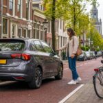 A person unlocking a car parked on a Dutch city street with bicycles nearby and traditional buildings in the background.