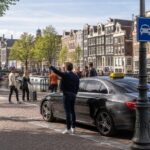 A taxi car parked near a canal in Amsterdam with people walking and cyclists passing by on a clear day.