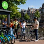 People renting bicycles near a bike rental kiosk on a street with traditional Amsterdam canal houses and a canal in the background.