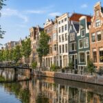 Row of traditional Dutch canal houses along a calm canal with bicycles parked nearby and a small pedestrian bridge.