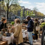 People selling and buying goods at an outdoor market in the Netherlands with traditional Dutch buildings and bicycles in the background.