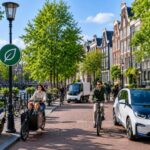 A clean Amsterdam street with bicycles, electric vehicles, and people walking near canal houses under a clear blue sky.