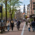 A busy street in the Netherlands with people walking and cycling, showing buildings and parked bicycles but no public toilets.