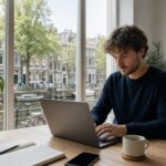 A young freelancer working on a laptop in a bright home office with a view of a Dutch canal, bicycles, and traditional buildings outside the window.