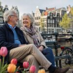 An elderly Dutch couple sitting on a bench near a canal with tulips and traditional houses in the background, smiling and enjoying a peaceful moment outdoors.