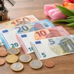 Euro banknotes and coins arranged on a wooden table with a small Dutch windmill figurine and a tulip flower in the background.