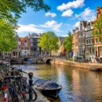 A scenic canal in Amsterdam with colorful narrow houses, bicycles parked along the water, a small boat on the canal, and a historic bridge in the background under a clear sky.