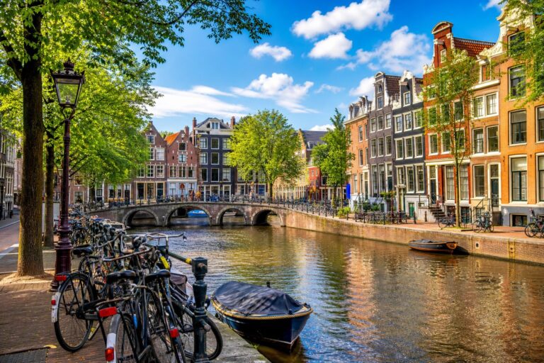 A scenic canal in Amsterdam with colorful narrow houses, bicycles parked along the water, a small boat on the canal, and a historic bridge in the background under a clear sky.