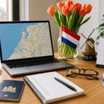 A workspace with a laptop showing a map, a passport, eyeglasses, a calendar, a cup of coffee, a small Dutch flag, tulips, and a windmill figurine, suggesting preparation for moving to the Netherlands.