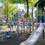 Children playing on colorful playground equipment in a green park with Amsterdam-style buildings and bicycles in the background.
