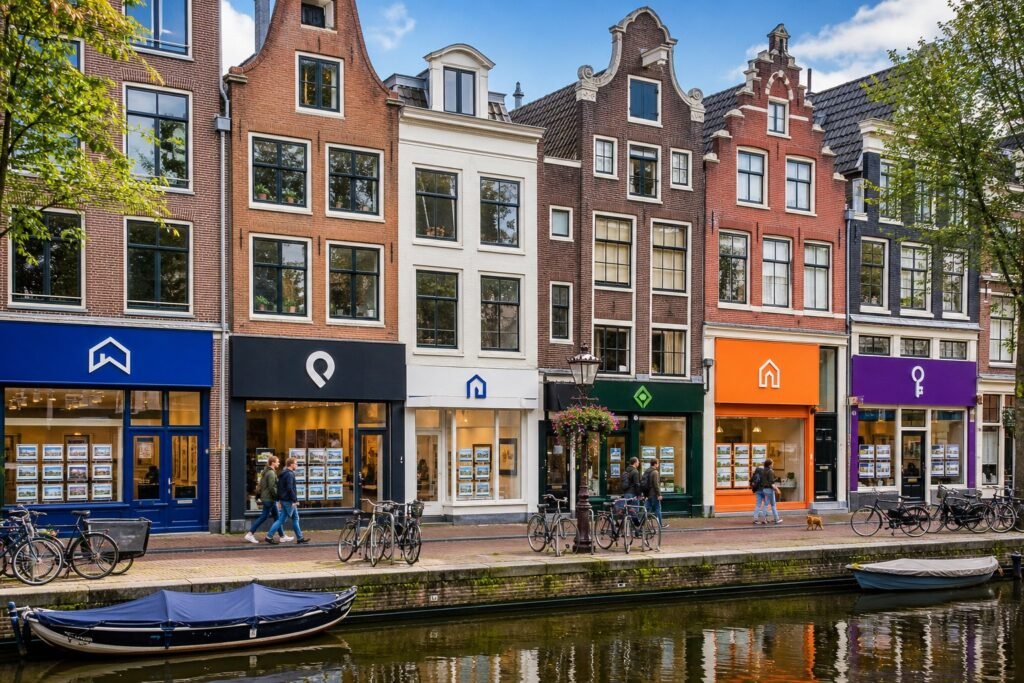 A canal-side street in Amsterdam with six rental agency storefronts, bicycles parked nearby, and classic Dutch buildings in the background.