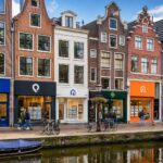 A canal-side street in Amsterdam with six rental agency storefronts, bicycles parked nearby, and classic Dutch buildings in the background.