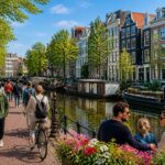 A sunny Amsterdam canal with colorful narrow houses, people walking and cycling along the water, and trees lining the street.