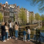 A sunny Amsterdam street with canal houses, bicycles, and people talking near a canal.