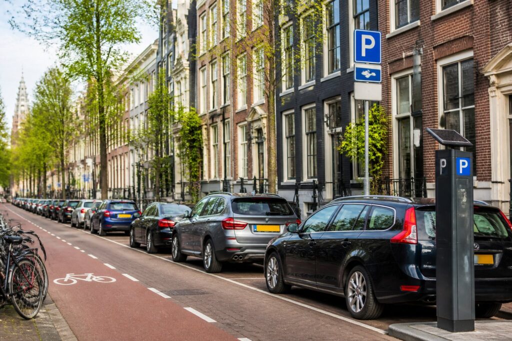 A quiet Amsterdam street with parked cars, bicycles, and traditional tall buildings along the sidewalk.