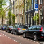 A quiet Amsterdam street with parked cars, bicycles, and traditional tall buildings along the sidewalk.