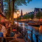 People socializing outdoors at canal-side cafes in Amsterdam with bicycles, boats, and traditional buildings in the background during early evening.