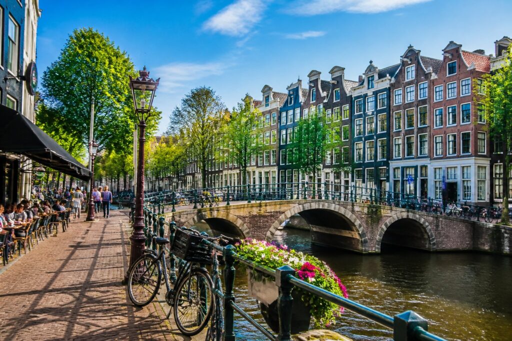 A sunny Amsterdam neighborhood with colorful canal houses, bicycles, a canal with a bridge, trees, and people walking or sitting at outdoor cafés.