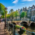 A sunny Amsterdam neighborhood with colorful canal houses, bicycles, a canal with a bridge, trees, and people walking or sitting at outdoor cafés.