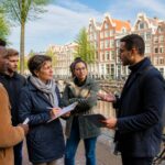A group of people discussing near Amsterdam canal houses with bicycles and water in the background.