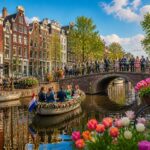 People celebrating on a cobblestone bridge and outdoor café terraces by a canal lined with colorful Amsterdam houses and blooming tulips.