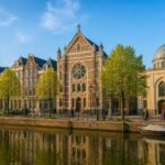 A canal in Amsterdam lined with historic buildings and diverse places of worship including a church, synagogue, and mosque under a clear sky.