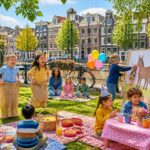 Children playing and celebrating at an outdoor party near a canal with Amsterdam buildings in the background.
