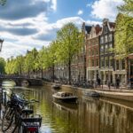 Canal in Amsterdam with traditional houses, people walking and cycling, partly cloudy sky, and calm water reflecting the scene.