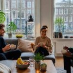 A bright living room in an Amsterdam apartment with young adults interacting and a view of canal houses outside.