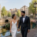 A couple walking hand-in-hand along an Amsterdam canal with traditional buildings and a bridge in the background.