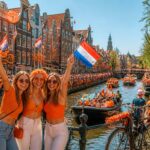 People celebrating Dutch festivities on a sunny street in Amsterdam with orange decorations, traditional buildings, bicycles, and boats along a canal.