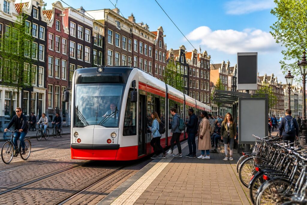 A busy Amsterdam tram moving along tracks with canal houses and people waiting at the tram stop, bicycles parked nearby.