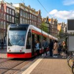 A busy Amsterdam tram moving along tracks with canal houses and people waiting at the tram stop, bicycles parked nearby.