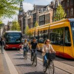A yellow and red tram and a city bus stopped on a cobblestone street in Amsterdam with canal houses and cyclists nearby.