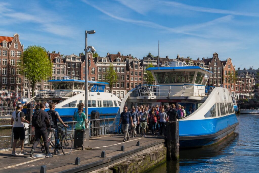 Ferries docked at an Amsterdam canal terminal with passengers boarding, surrounded by traditional canal houses and calm water.