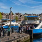 Ferries docked at an Amsterdam canal terminal with passengers boarding, surrounded by traditional canal houses and calm water.
