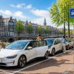People using electric cars parked by a canal with traditional Amsterdam buildings and bicycles nearby.