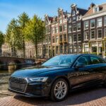 A modern car parked on a cobblestone street in Amsterdam near a canal with traditional buildings and bridges in the background.
