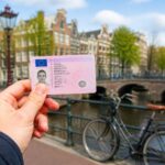 A person holding a European-style driver's license with blurred Amsterdam canal houses and bicycles in the background.