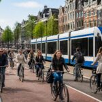 A busy Amsterdam street with cyclists on a bike lane, a tram passing by, and canal houses in the background.