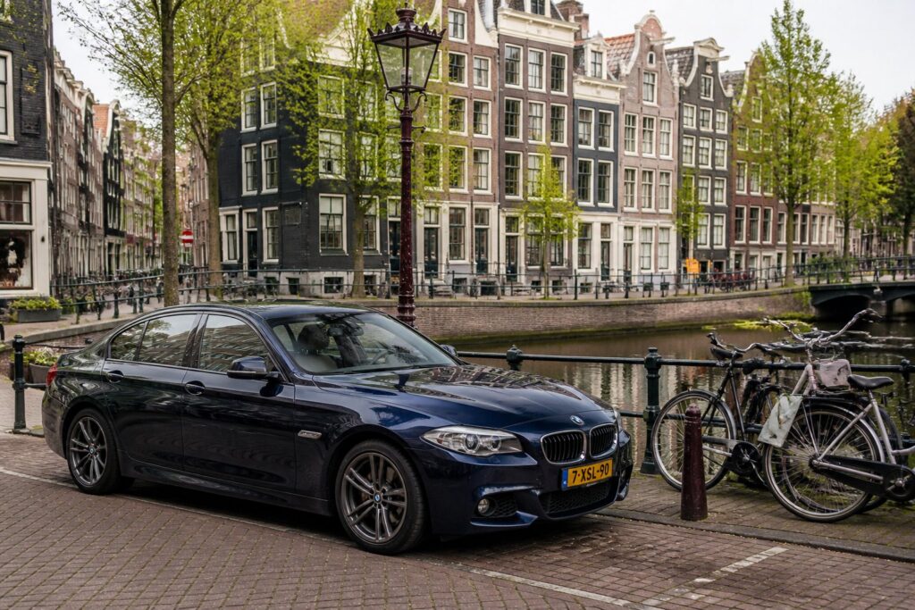 A modern car parked on a narrow street in Amsterdam with brick buildings, bicycles, and a canal nearby.