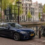 A modern car parked on a narrow street in Amsterdam with brick buildings, bicycles, and a canal nearby.
