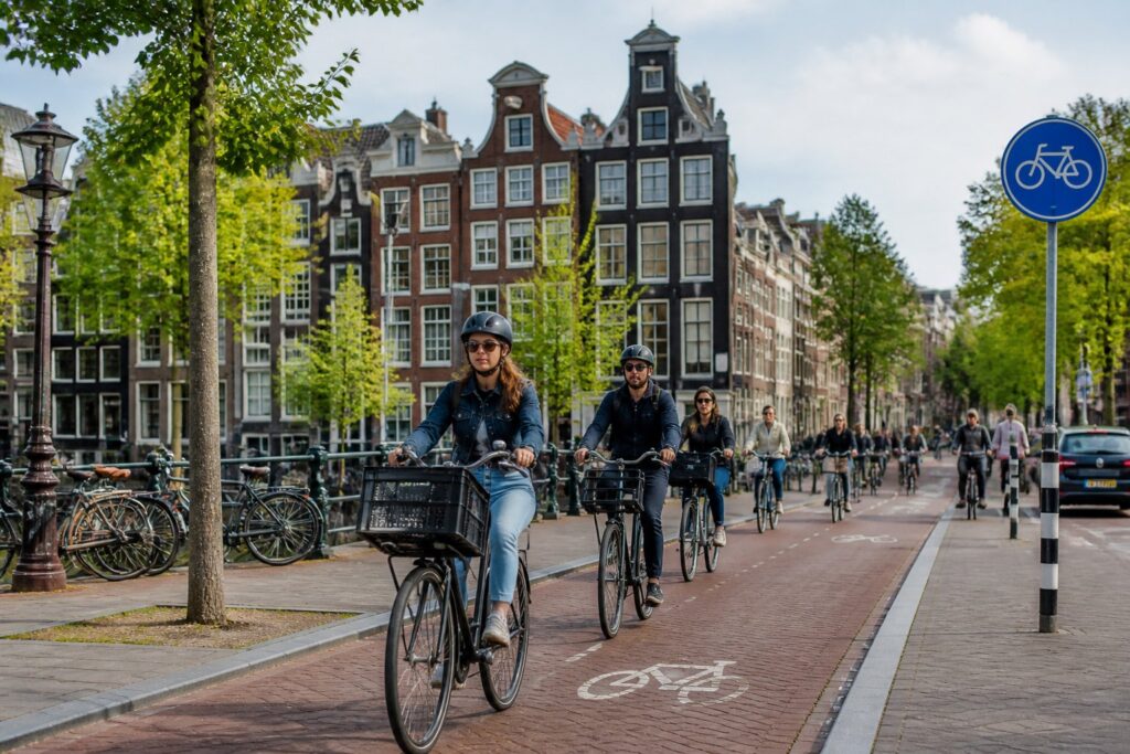 Cyclists riding on a dedicated bike lane in a busy Amsterdam street with canal houses in the background.