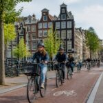 Cyclists riding on a dedicated bike lane in a busy Amsterdam street with canal houses in the background.