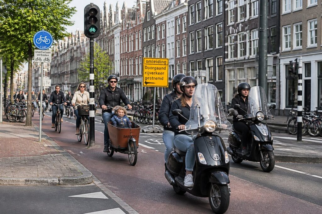 A busy Amsterdam street with bicycles and mopeds riding in designated lanes alongside traditional canal houses.