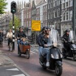 A busy Amsterdam street with bicycles and mopeds riding in designated lanes alongside traditional canal houses.