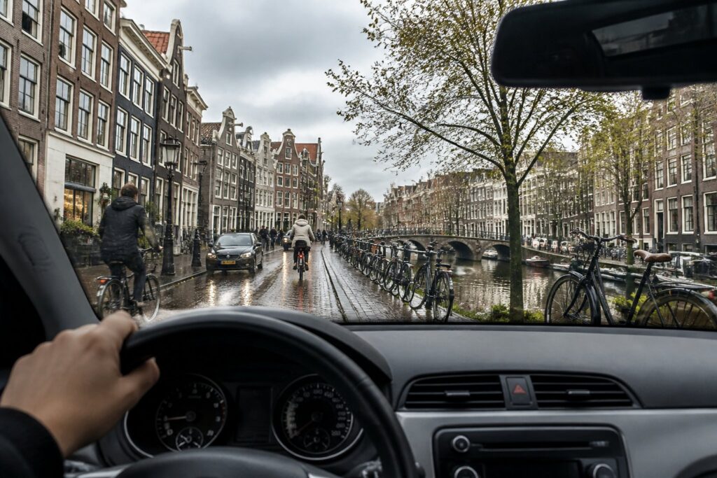 View from inside a car driving through a street in Amsterdam with canal houses, bicycles, and a canal in the background.